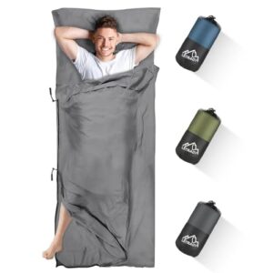 Junger Mann liegt entspannt in einem grauen Hüttenschlafsack aus Mikrofaser, ideal für Backpacking und Hostelaufenthalte, umgeben von drei kompakten Schlafsäcken in Blau, Grün und Schwarz.