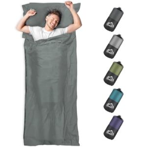 Mann entspannt in einem dunkelgrauen Hüttenschlafsack aus Mikrofaser, ideal für Backpacking und Hostels, mit einem weiteren Set kleiner Schlafsäcke in verschiedenen Farben im Hintergrund