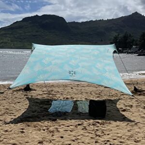 Neso Grande Beach Zelt in leicht transparentem Aquaton, das Schatten am Strand bietet. Hintergründe mit grünem Berg und ruhigem Gewässer.